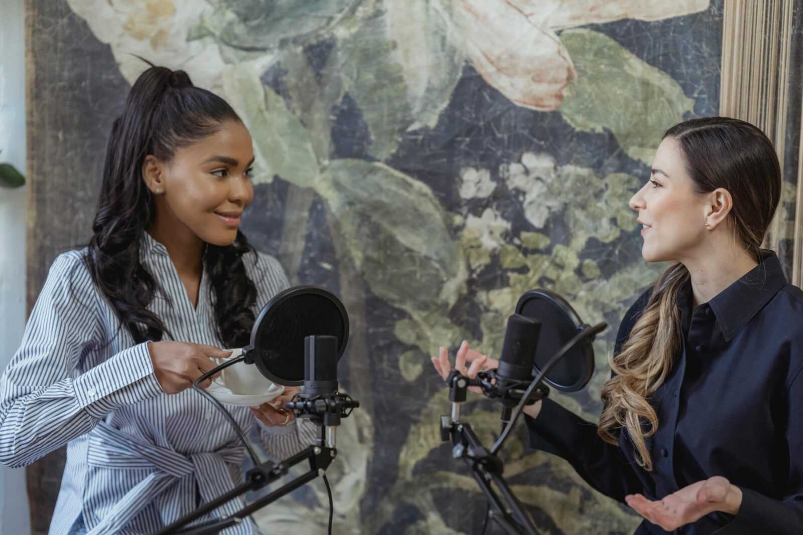 Two women hosting a podcast, engaged in conversation with microphones indoors.