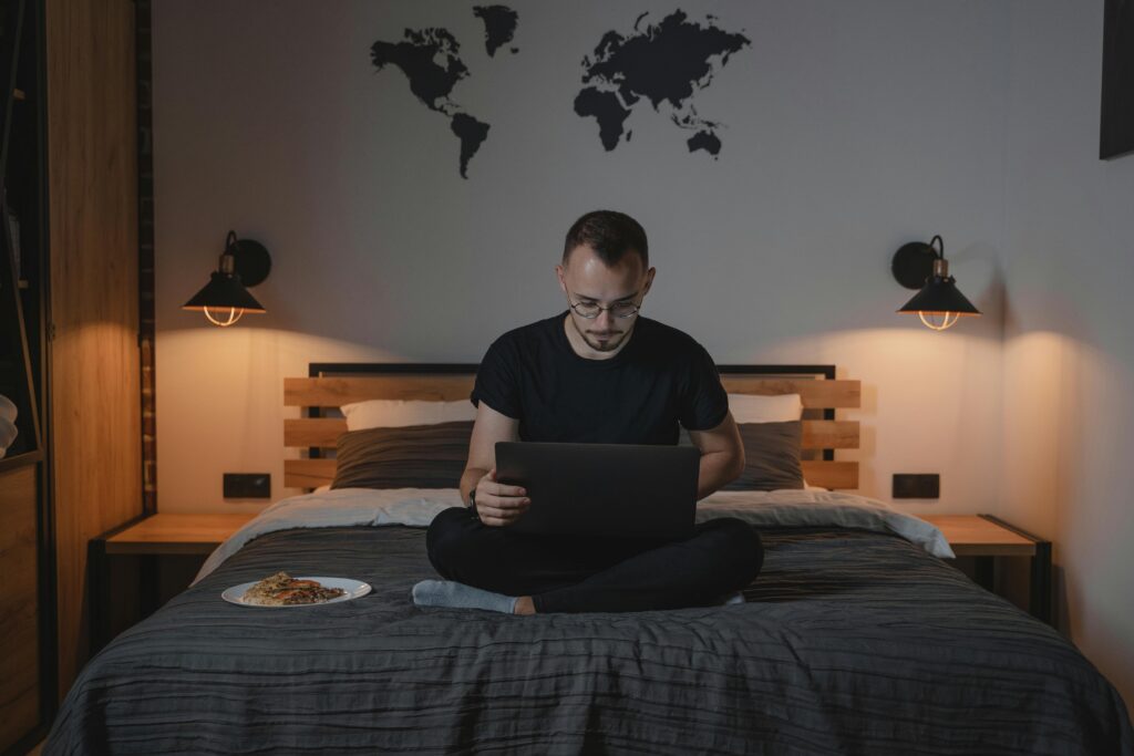 Man working on a laptop in a cozy bedroom setting, showcasing remote work lifestyle.