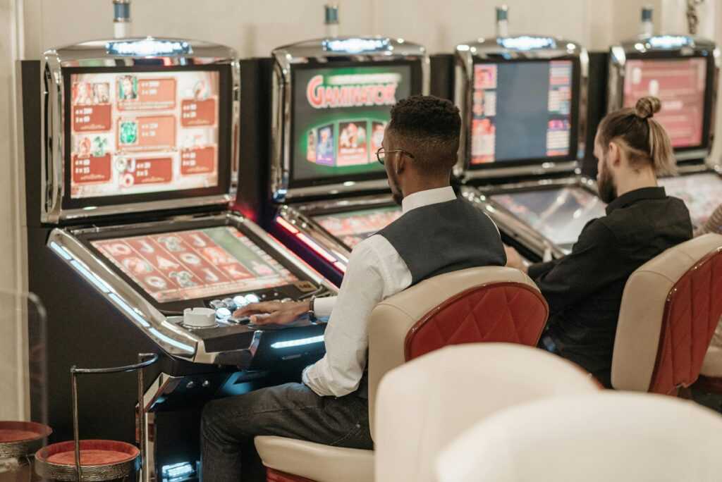 Two men engaged in gaming at a casino, seated at slot machines indoors.